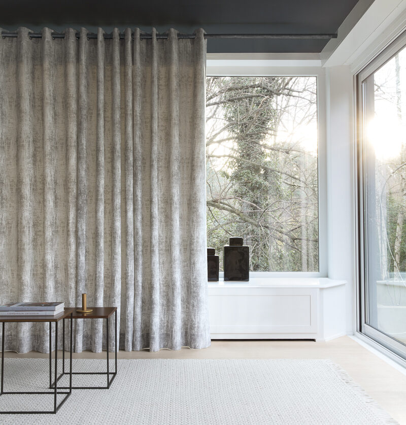 Modern living room corner with large windows featuring Denver-inspired window treatments, grey curtains, and minimalistic decor, overlooking trees.
