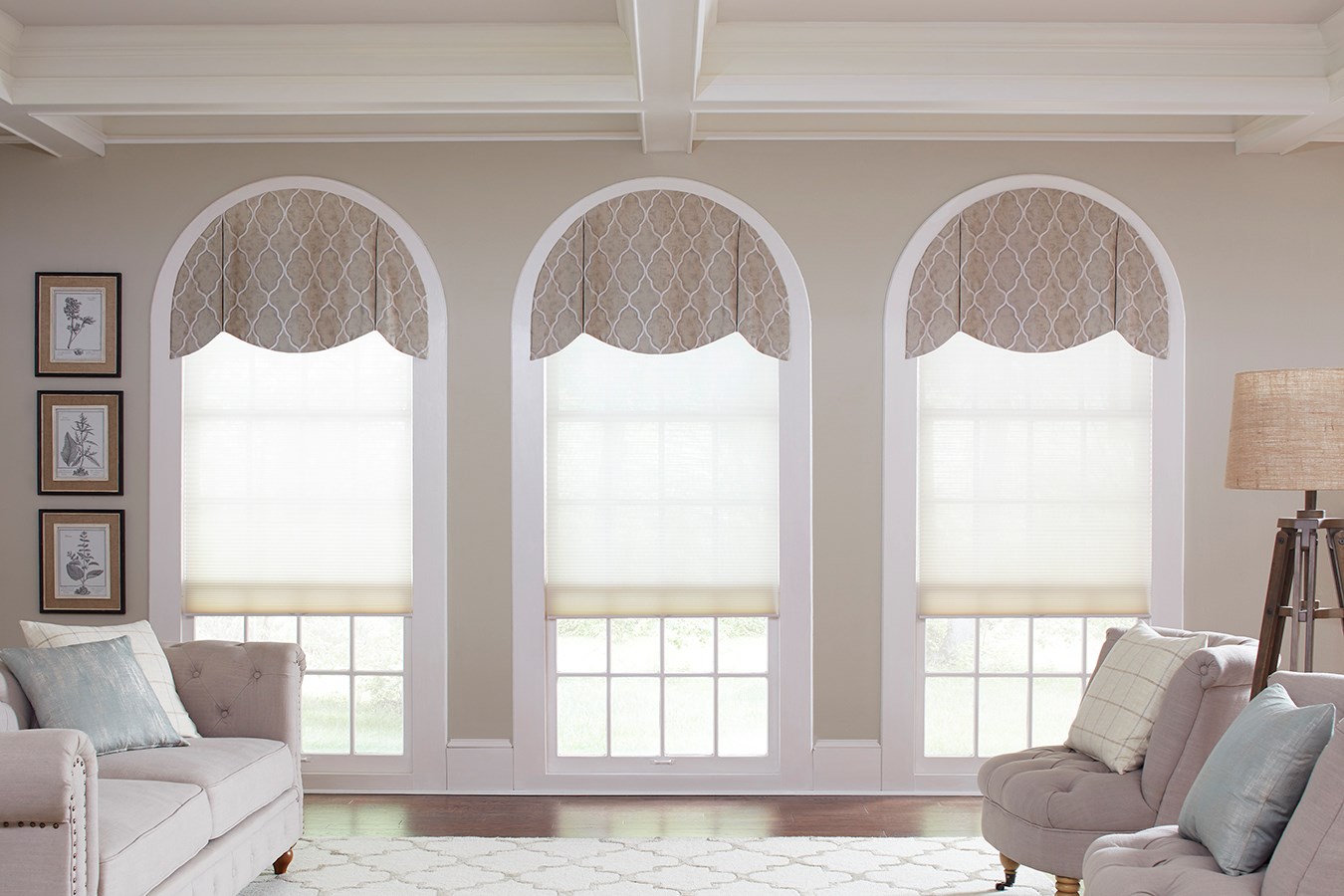 Three large windows with patterned half-circle valances and sheer roller shades, offering an elegant window treatment in a stylish living room with neutral-toned furniture and framed artwork.