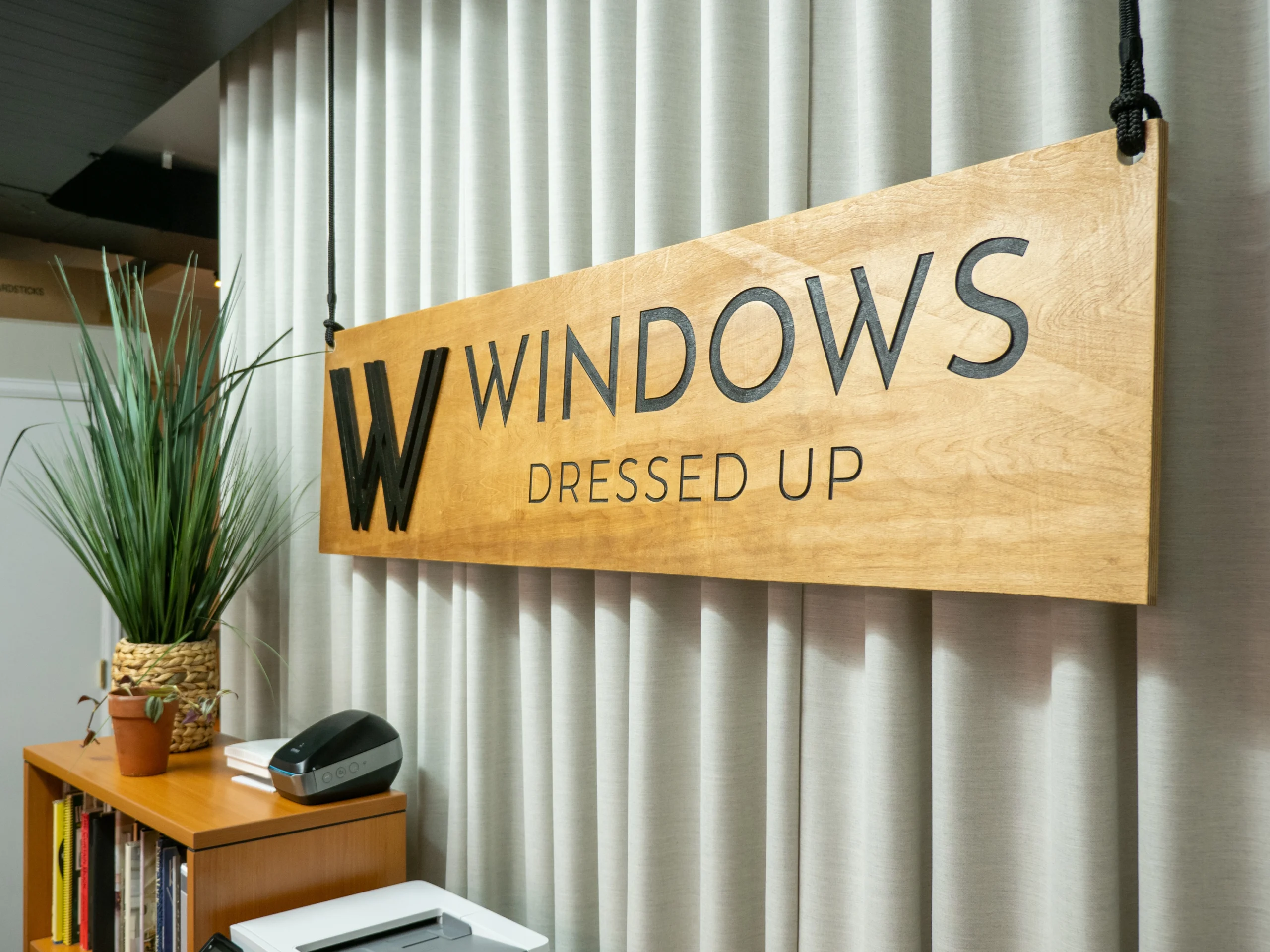 Wooden sign reading "windows dressed up" hanging in an office, with a potted plant and drapery in the background.