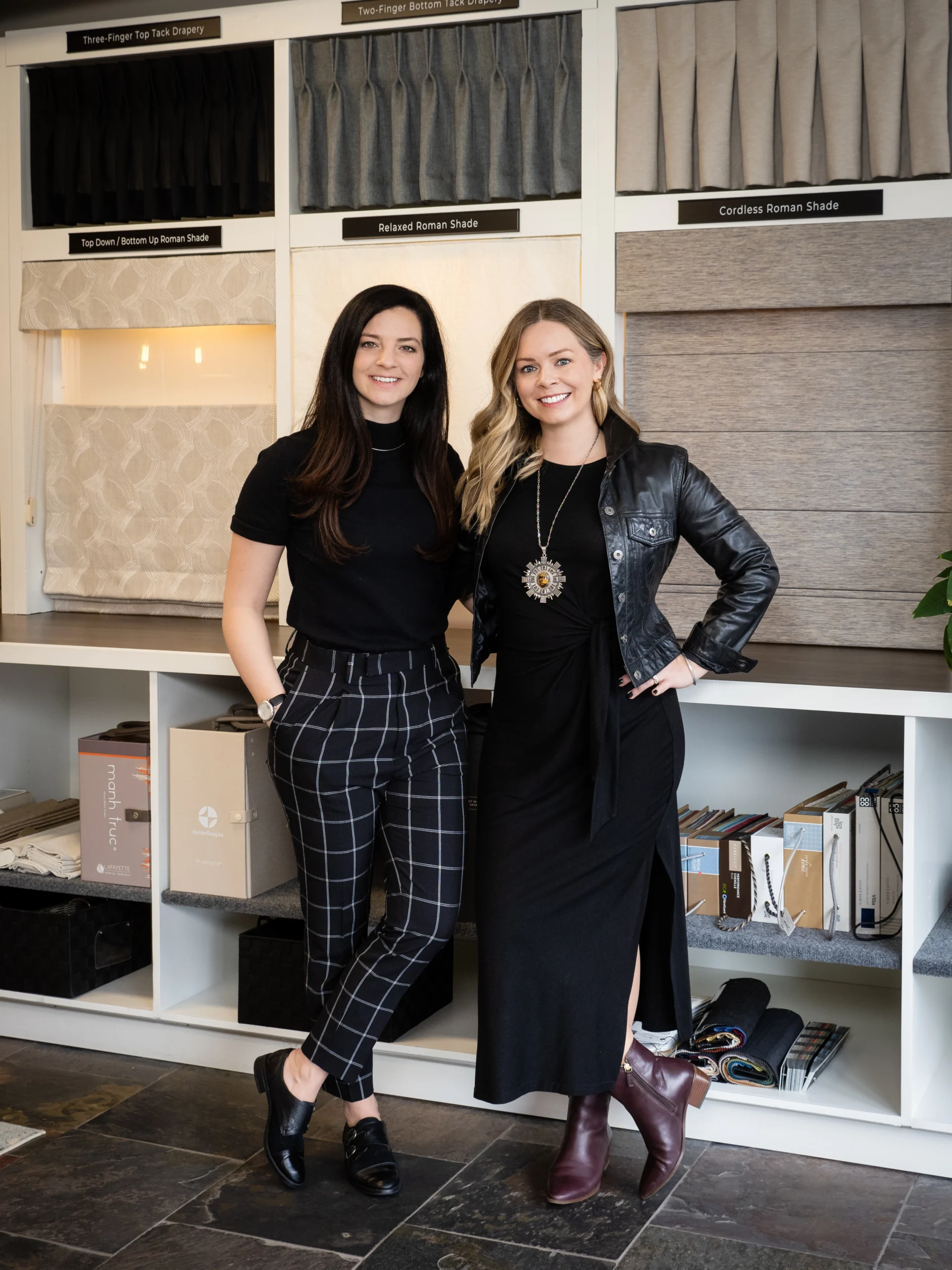 Two women standing in a Colorado showroom with various blinds and window treatment samples displayed behind them. One wears a black outfit, and the other a black dress with a leather jacket.