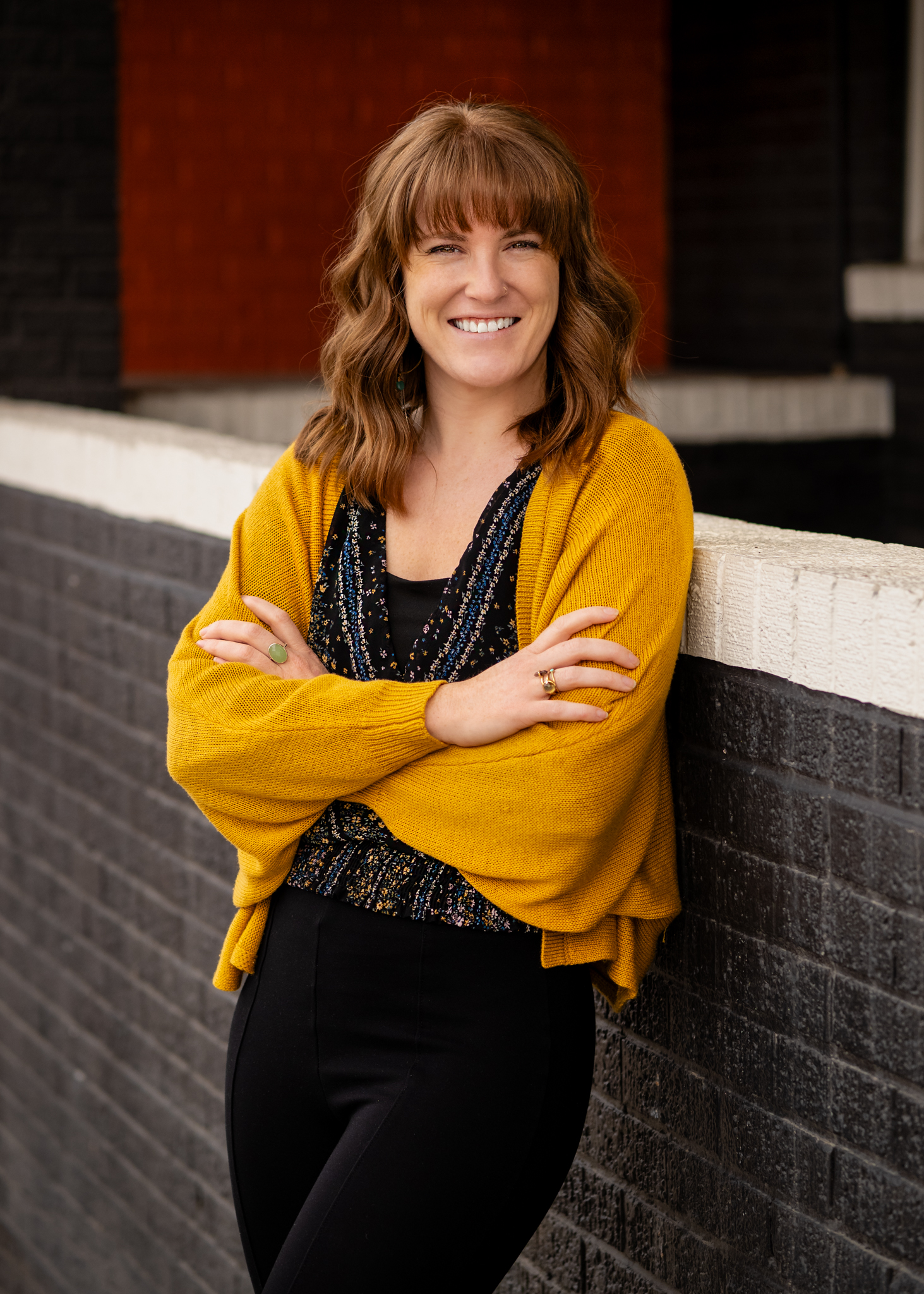 A woman smiling, standing with arms crossed, dressed up in a yellow cardigan and black pants, leaning against a brick wall.