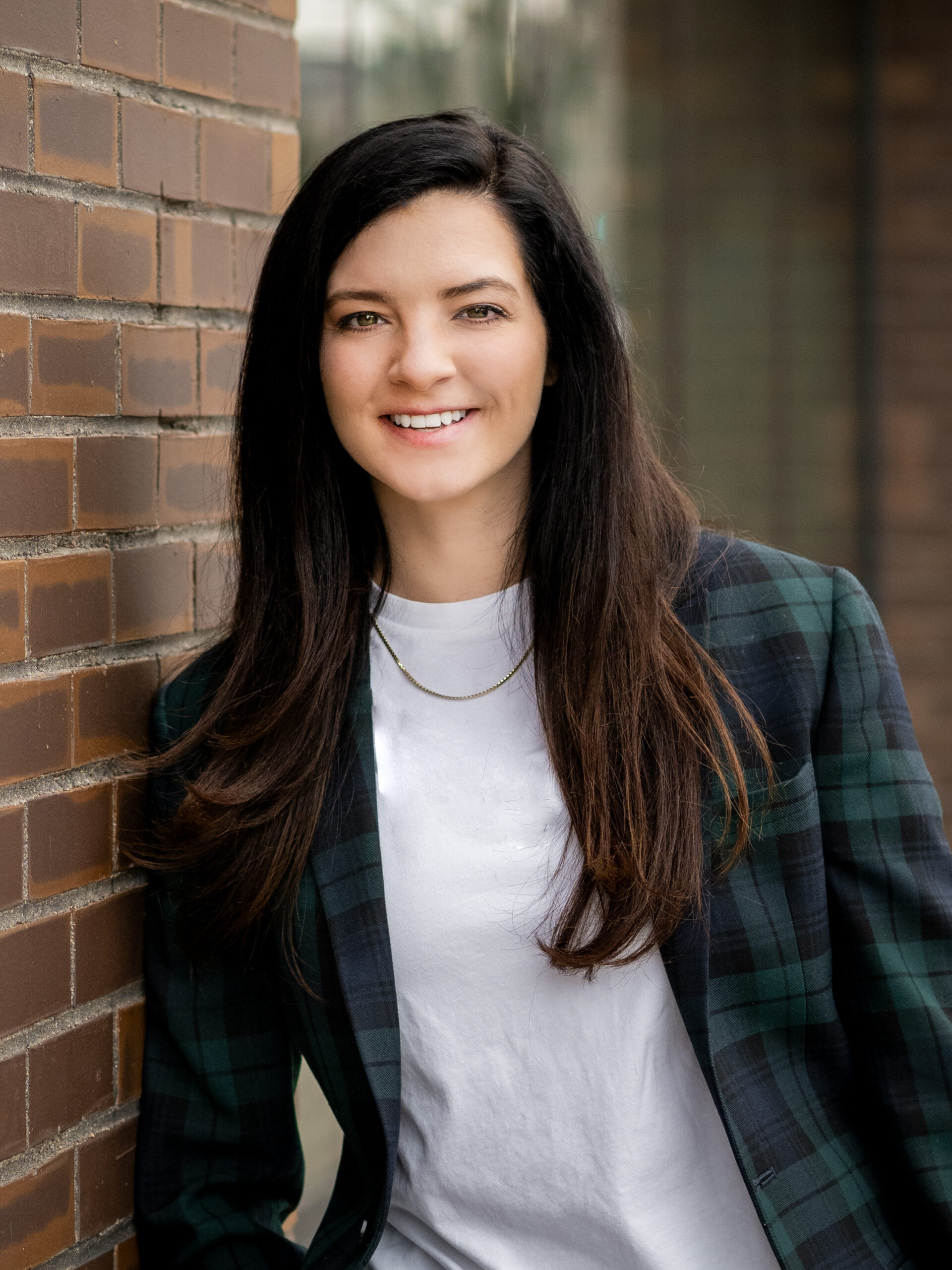 A woman with long dark hair, dressed up in a plaid blazer and white shirt, smiling while standing against a brick wall.