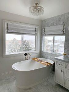 Luxurious bathroom with a freestanding tub, marble tiles, crystal chandelier, and two windows with winter view.