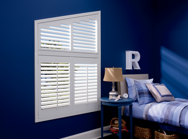 A stylishly arranged bedroom featuring windows dressed up with white shutters, blue walls, and bedding matching with a bedside lamp and decorative letter 'r'.