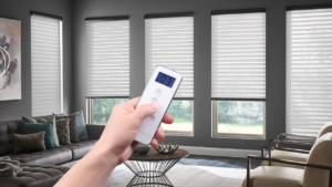 A hand holding a remote control to adjust settings in a modern living room with stylish blinds and furniture.