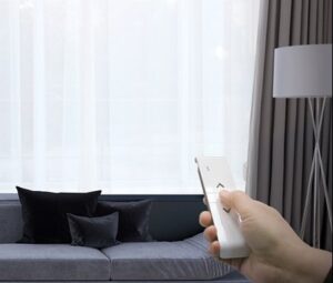 A person's hand holding a remote control to adjust the curtains in a modern living room with a grey sofa and white lamp.