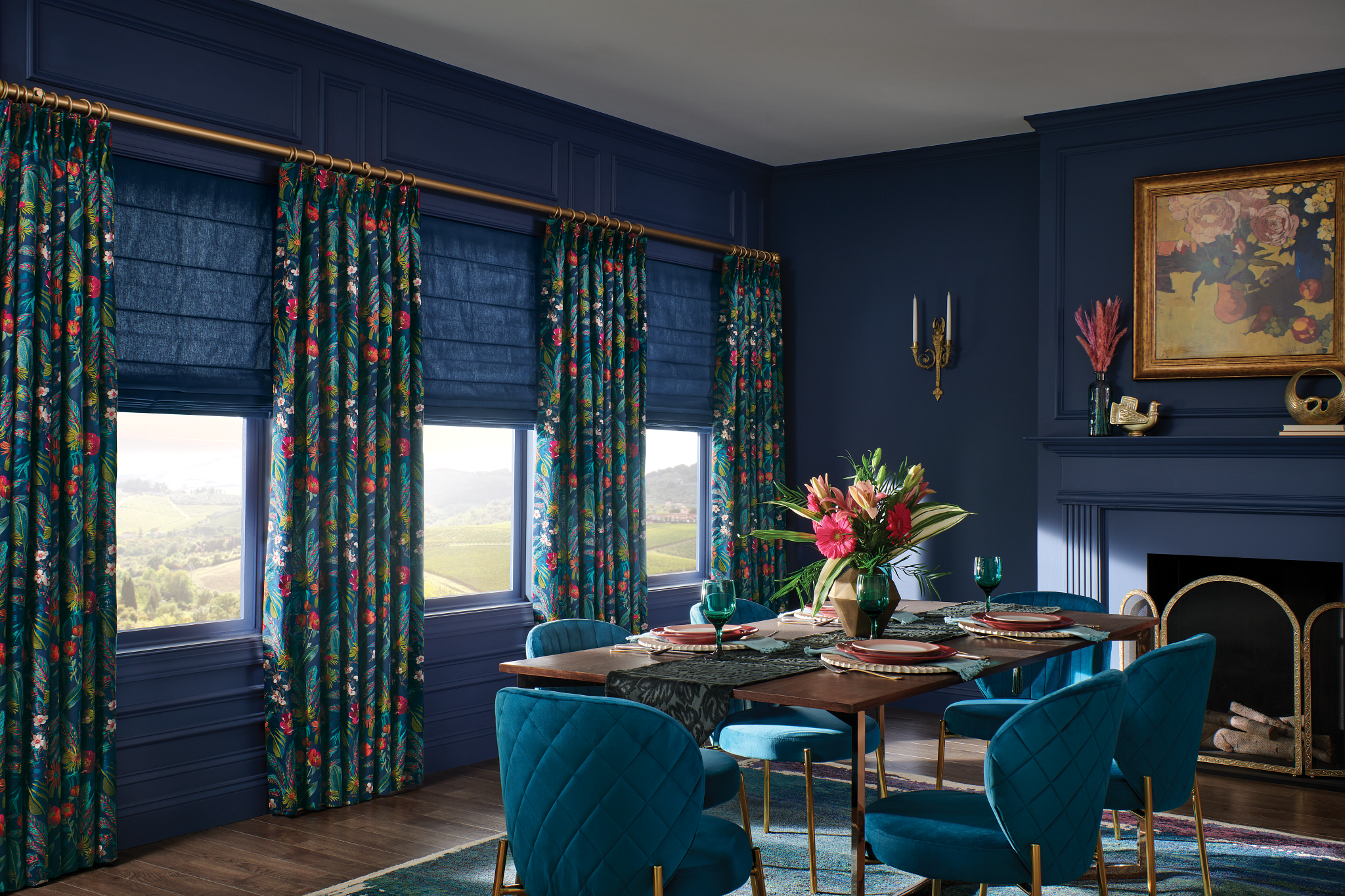 A dining room with blue walls, blue floral-patterned curtains from Windows Dressed Up Design Studio, and a wooden table set for six. A vase with flowers graces the table, while a painting and sconces adorn the walls.