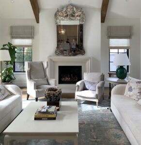 A living room with a fireplace, two armchairs, two sofas, a coffee table with books, table lamps, plants, and a large ornate mirror above the mantel.