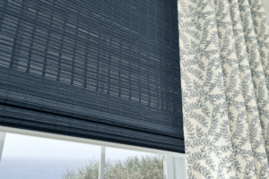 Close-up of a window with dark blue bamboo blinds and white curtains with a blue leafy pattern. The view outside shows trees and a distant body of water.