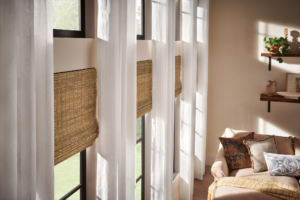 A sunlit room with tall windows featuring sheer white curtains and woven blinds. A beige sofa with assorted pillows is to the right, and wall-mounted shelves hold various decor items.