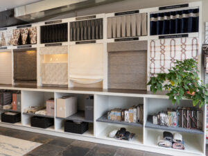 Display of various window treatments and fabric samples in a showroom, with labeled sections and books on shelves below.