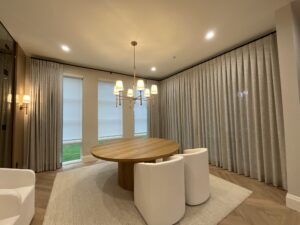 A modern dining room with a round wooden table, four white chairs, a chandelier, and large windows with curtains.