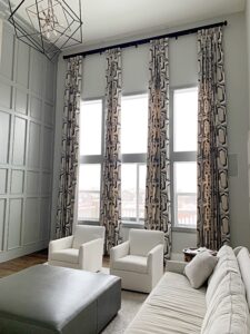 A modern living room with tall patterned curtains, white walls, a grey ottoman, and two white armchairs beside a light-colored sofa. A geometric light fixture hangs from the ceiling.