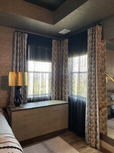 A cozy room corner features two windows with patterned curtains and sheer drapes. A wooden dresser sits below with a brass lamp on top, and a sofa edge is visible on the left.