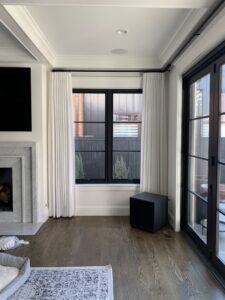 A modern living room with large windows, white curtains, and a wall-mounted TV. There is a fireplace on the left and a dark wooden floor with a rug.