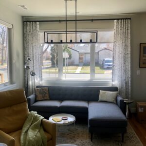 A cozy living room with a gray sectional sofa, pillows, and a yellow armchair. A modern chandelier hangs above, and large windows overlook a suburban street. A round table holds books and a plant.