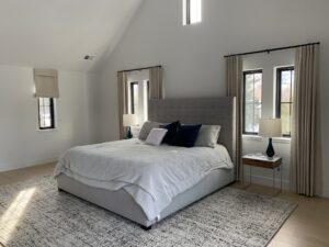 A bedroom with a gray upholstered bed, white bedding, navy throw pillows, two side tables with lamps, and windows with beige curtains. A textured gray rug covers the light wood floor.