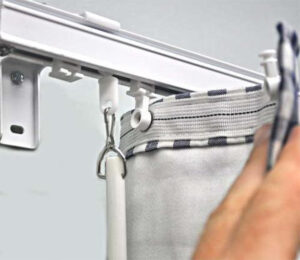 Close-up of a hand attaching a curtain to a rail with hooks. The curtain has a striped top and is being fastened to a white overhead track.