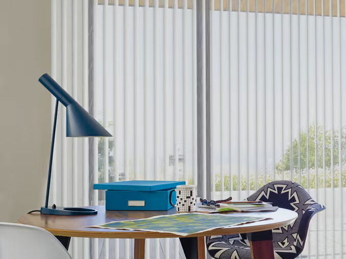A round wooden desk with books, a lamp, and blue stationery sits in front of large windows covered by crisp white Hunter Douglas vertical blinds. Two modern chairs are arranged around the desk.