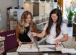 Two women review fabric samples and color swatches in a showroom, standing at a counter with display binders and material samples visible in the background.