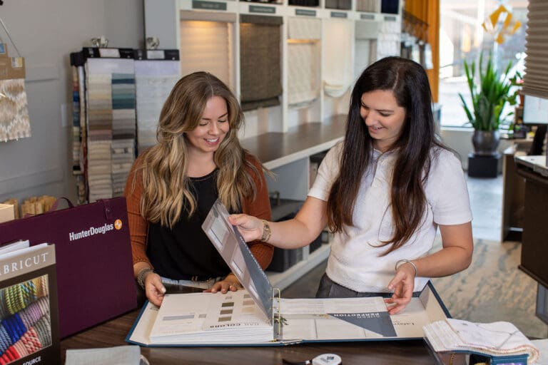 Two women review fabric samples and color swatches in a showroom, standing at a counter with display binders and material samples visible in the background.