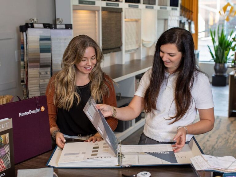 Two women review fabric samples and color swatches in a showroom, standing at a counter with display binders and material samples visible in the background.