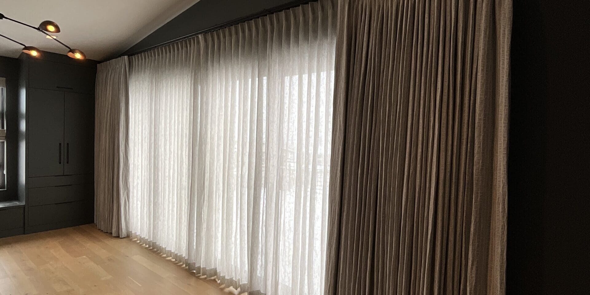 A modern room with light wooden floors, dark walls, and large windows covered by sheer white curtains and heavier beige drapes.