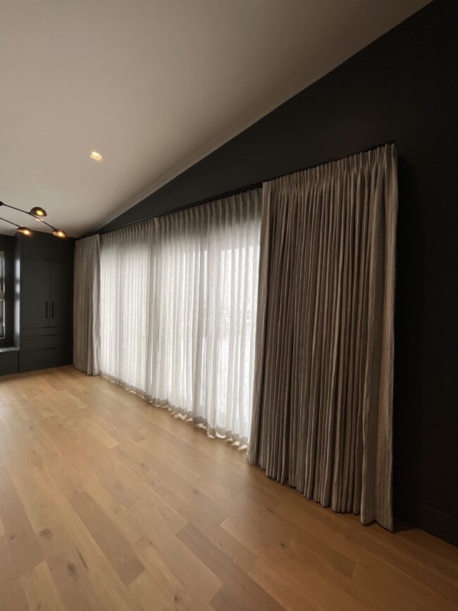 IMG_1024 A modern room with light wooden floors, dark walls, and large windows covered by sheer white curtains and heavier beige drapes.