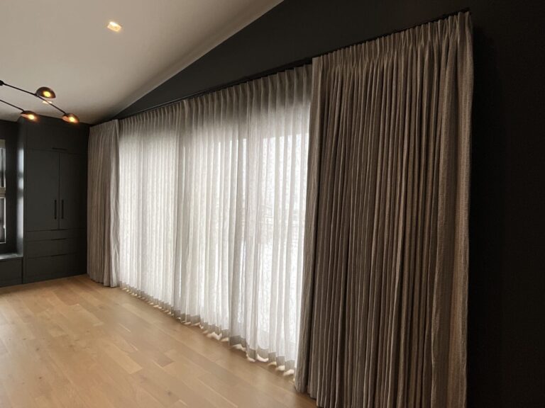 A modern room with light wooden floors, dark walls, and large windows covered by sheer white curtains and heavier beige drapes.