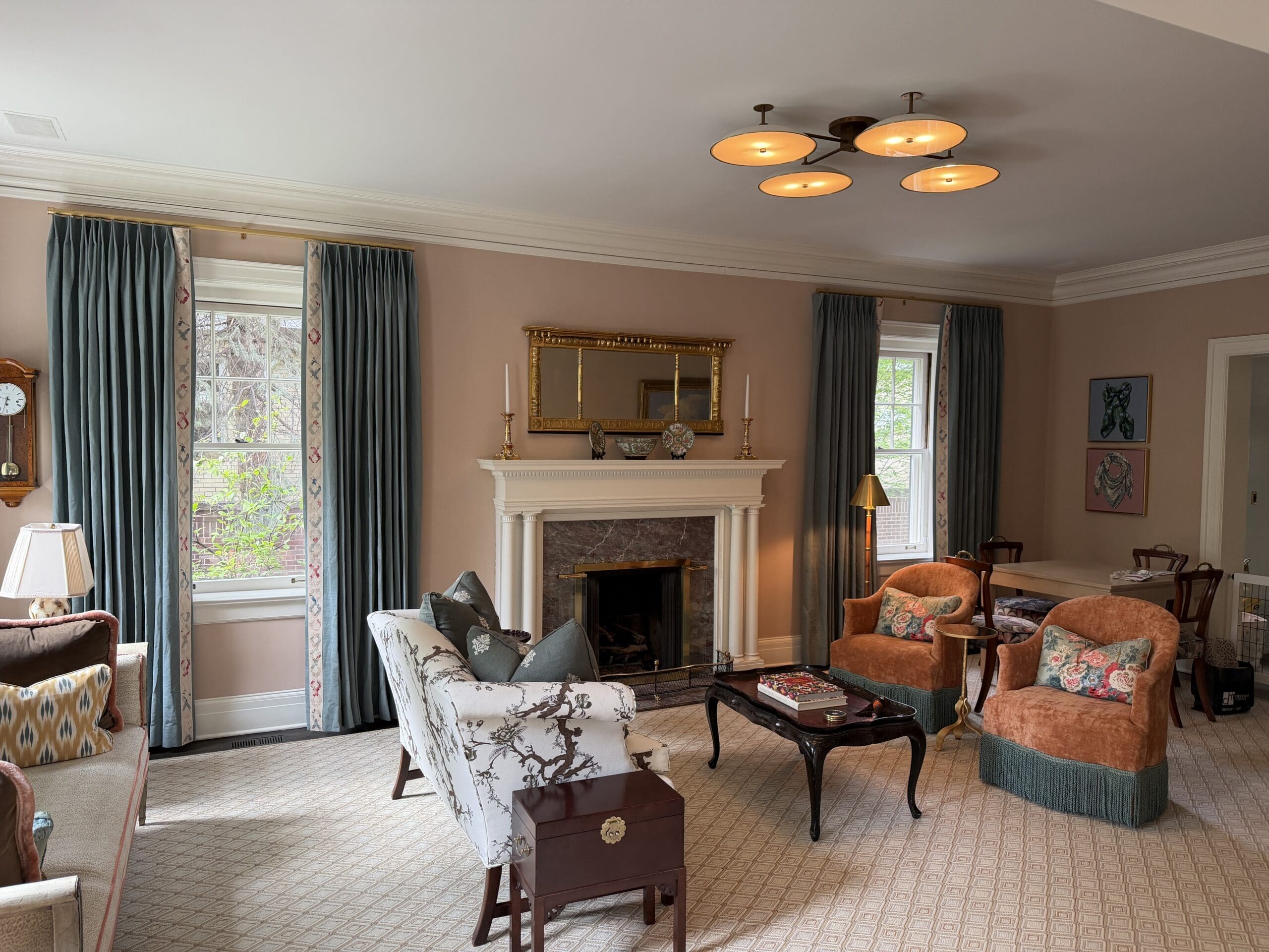 heirloom quality drapery A traditional living room with patterned chairs, a sofa, a fireplace with a mirror above, blue curtains, and framed art on beige walls. A coffee table sits in the center of the room.