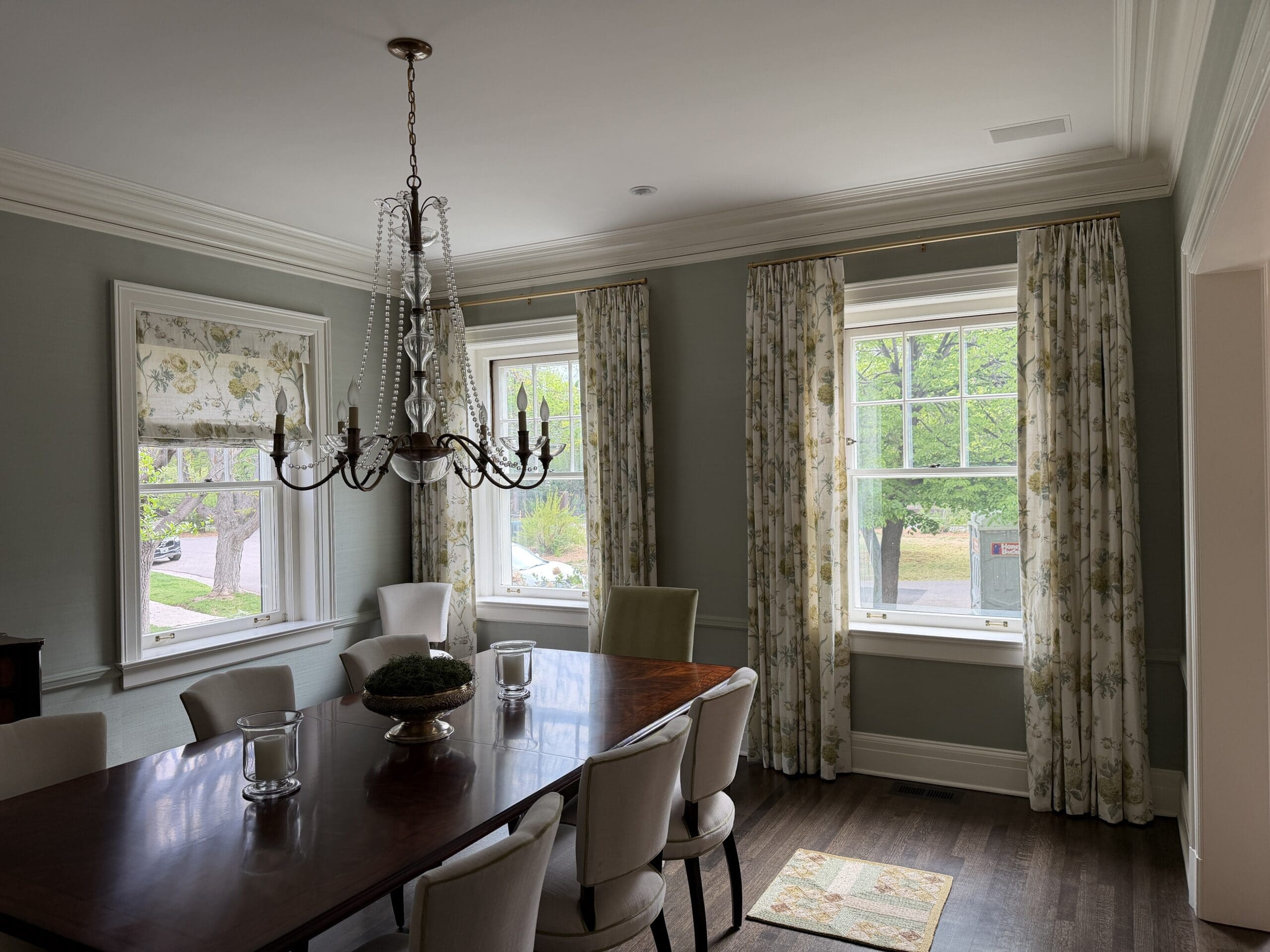 IMG_3218 A dining room with a wooden table, eight chairs, a chandelier, floral curtains, and three large windows letting in natural light.