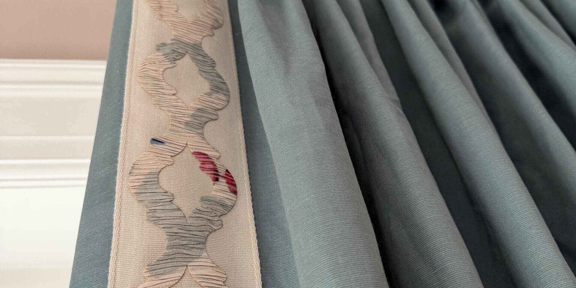 custom drapery design denver Close-up of blue pleated curtains hanging from a gold curtain rod, with a decorative cream and pink embroidered trim along one edge.