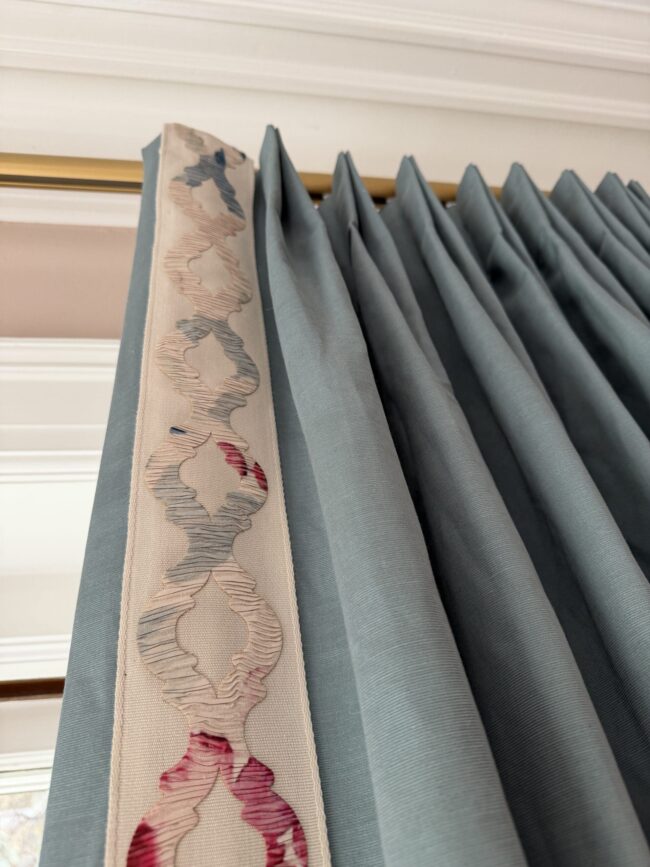 Close-up of blue pleated curtains hanging from a gold curtain rod, with a decorative cream and pink embroidered trim along one edge.