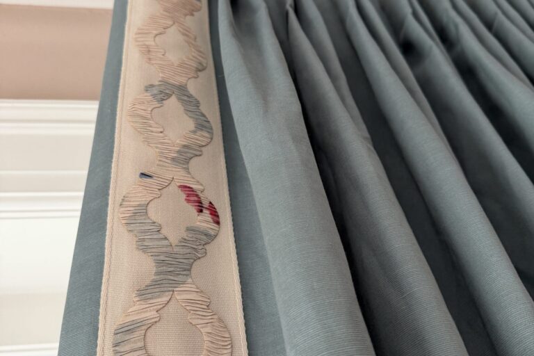 Close-up of blue pleated curtains hanging from a gold curtain rod, with a decorative cream and pink embroidered trim along one edge.