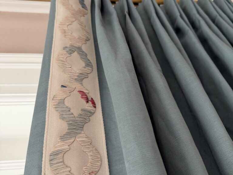 Close-up of blue pleated curtains hanging from a gold curtain rod, with a decorative cream and pink embroidered trim along one edge.