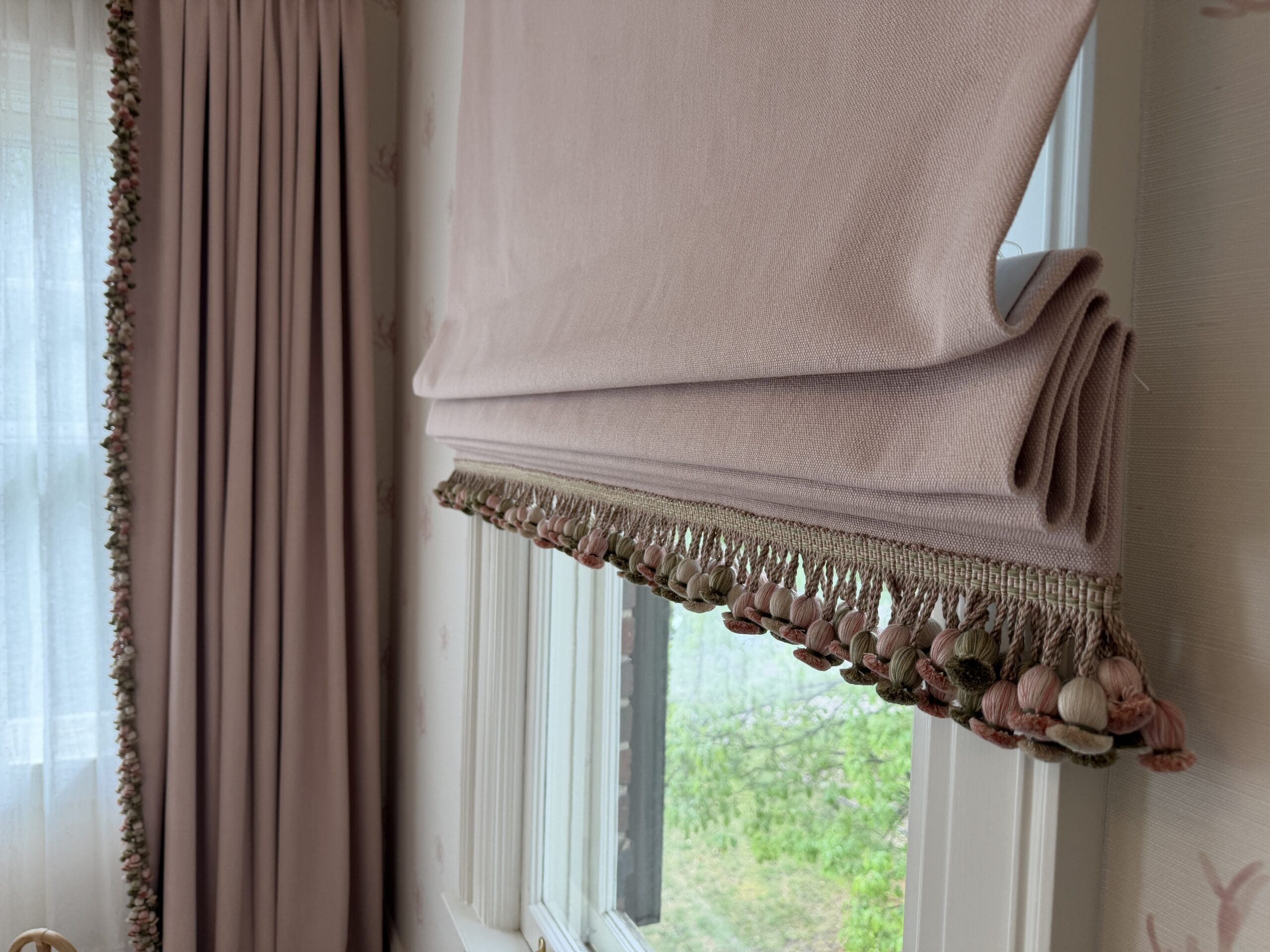 bespoke roman shades denver A close-up of a pink Roman shade with decorative tassels, partially raised on a window next to matching pink curtains in a softly lit room.