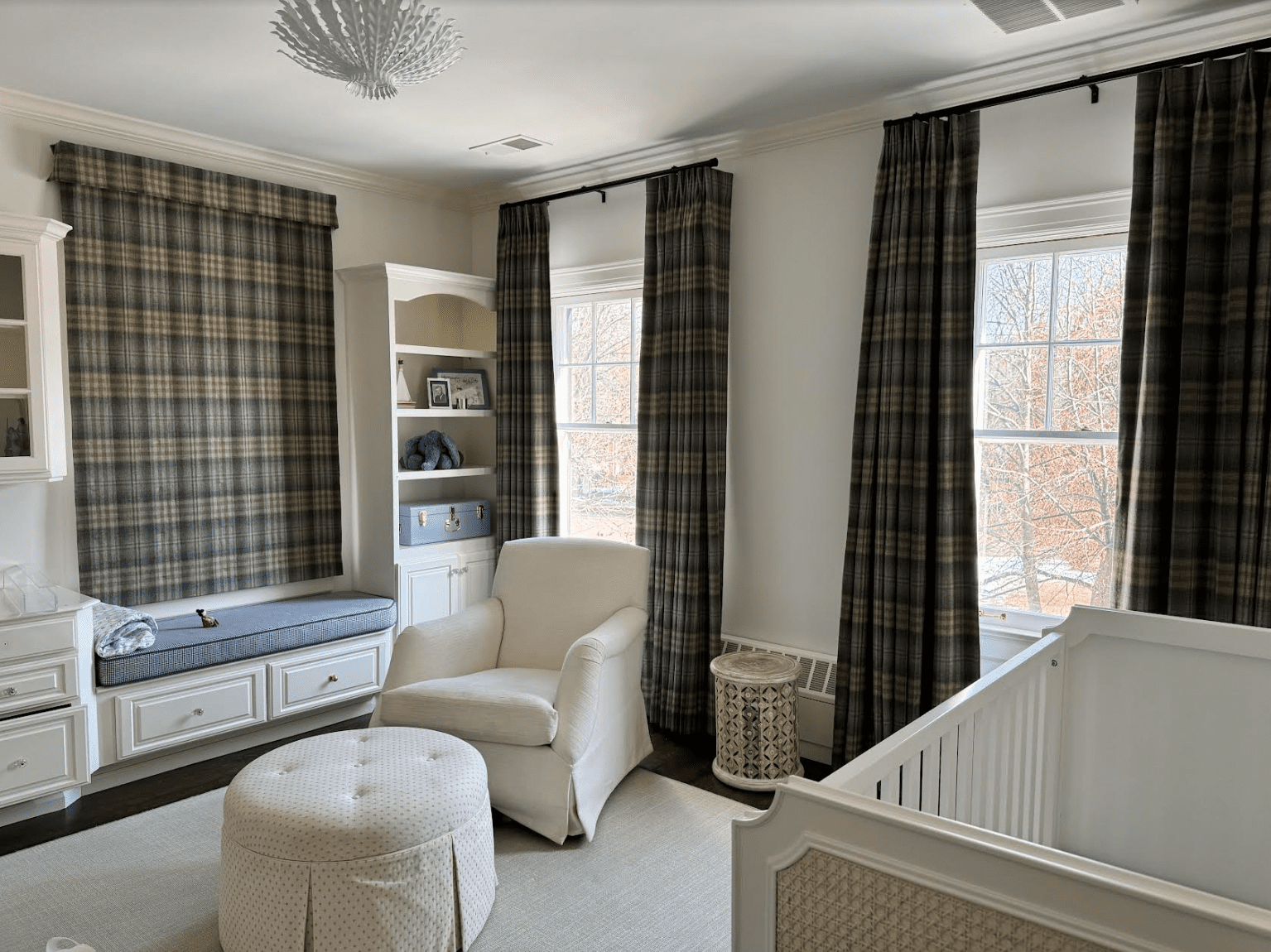 custom drapery denver A nursery with a white crib, cream armchair, plaid curtains, built-in shelves, window seat, and ottoman, with sunlight streaming through two windows.