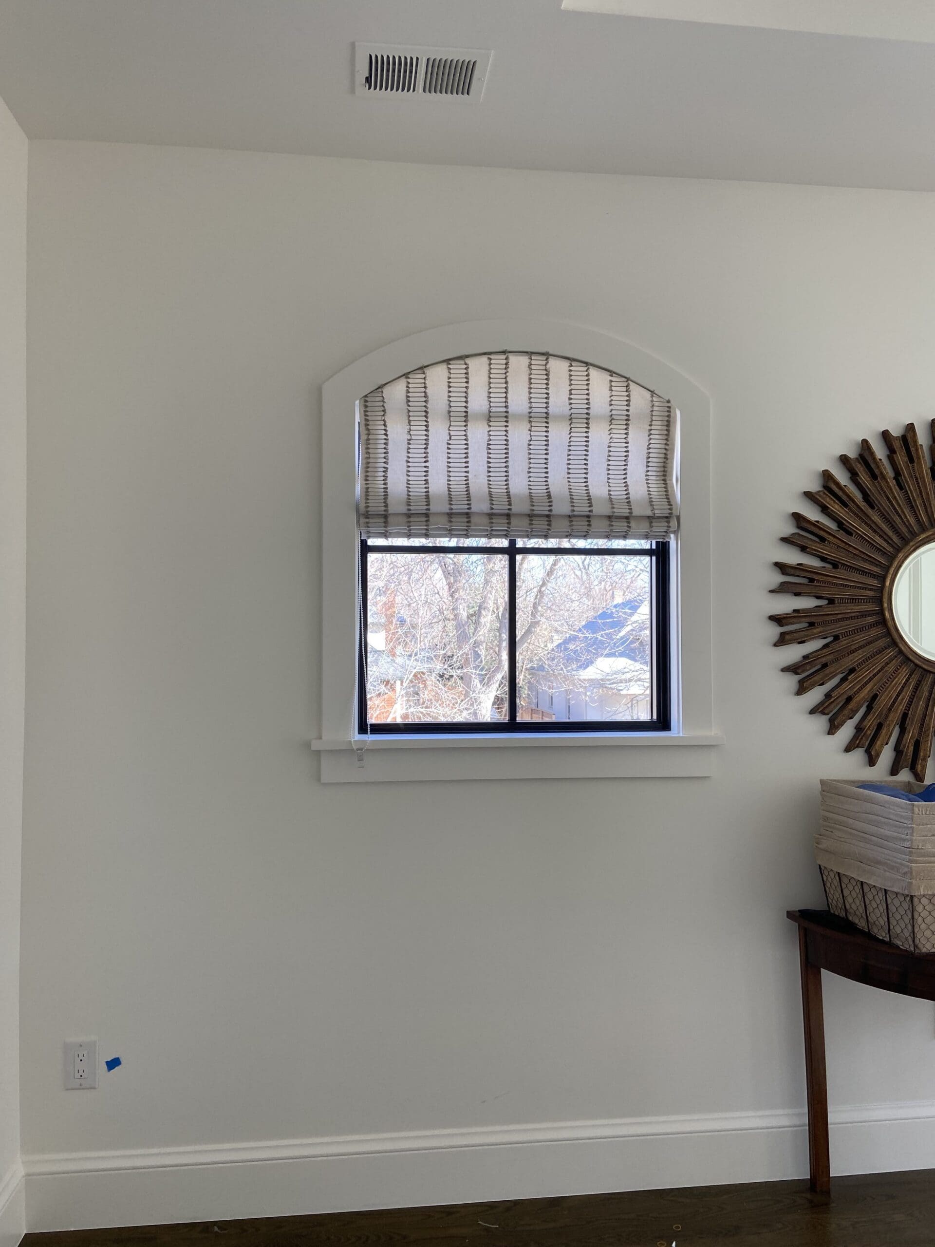 arched-window-custom-roman-shade-denver.jpg A window with a striped Roman shade partially open, revealing a view of trees outside. A round mirror and stacked boxes sit on a wooden table to the right.