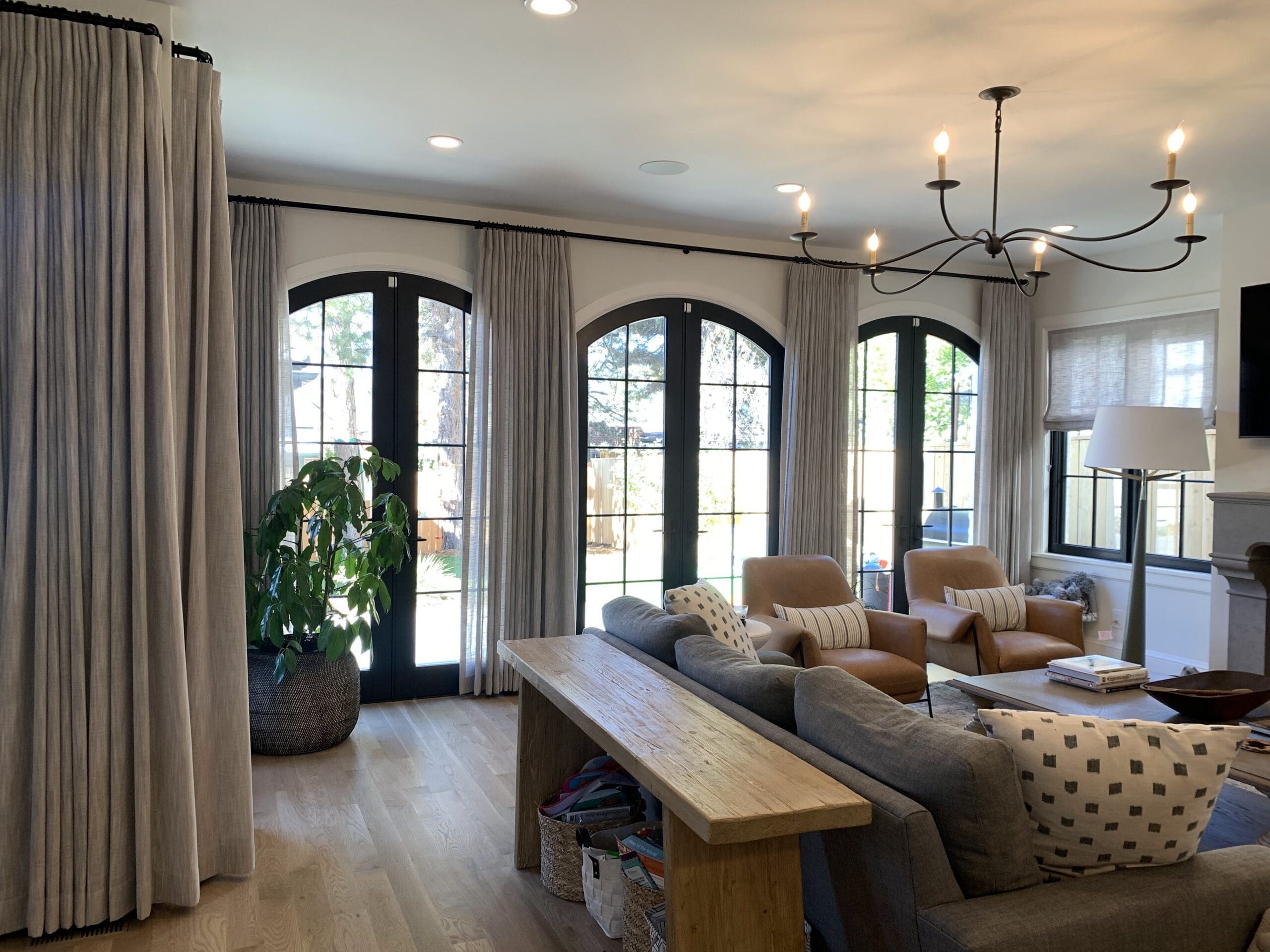 custom-drapery-sliding-patio-doors-denver-home.jpg Living room with a gray sofa, two tan chairs, a wooden bench, a potted plant, and large arched black-framed windows with light curtains.