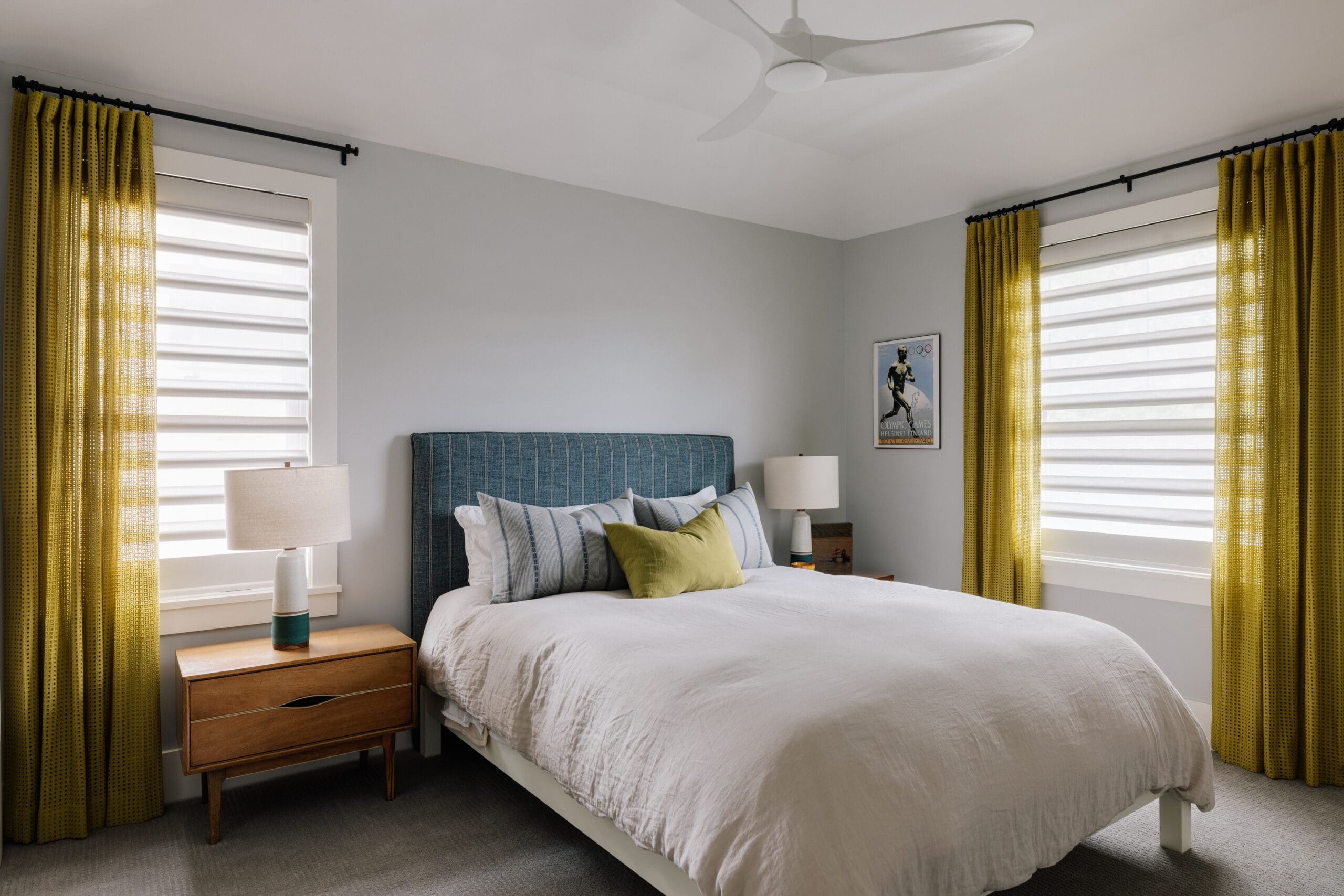 Windows Dressed Up | August Photoshoots A modern bedroom with a blue upholstered bed, white bedding, two wooden nightstands with lamps, yellow curtains, and a poster on the wall.