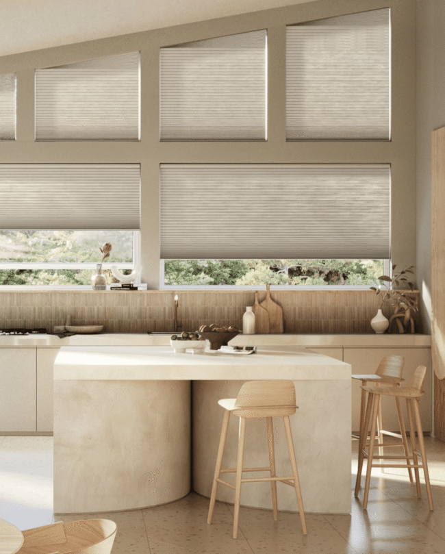 Modern kitchen with light wood cabinets, a curved island with two wooden stools, and large windows with beige blinds letting in natural light.