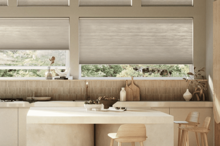 Modern kitchen with light wood cabinets, a curved island with two wooden stools, and large windows with beige blinds letting in natural light.