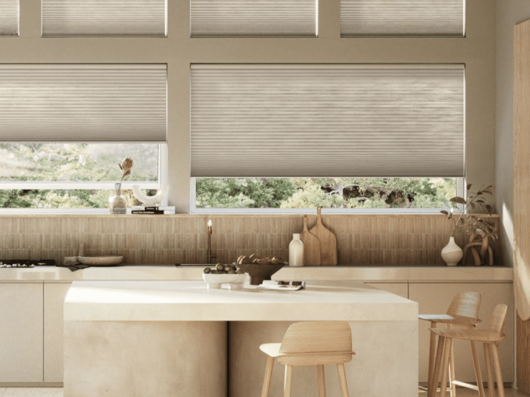 Modern kitchen with light wood cabinets, a curved island with two wooden stools, and large windows with beige blinds letting in natural light.