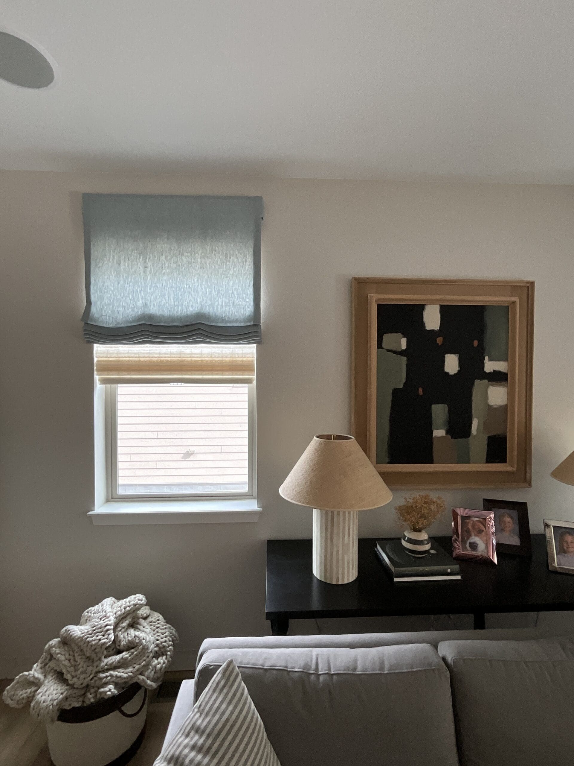 motorized-roman-shades-denver.jpg A living room with a window featuring two shades, a black console table with lamps, framed photos, a basket with a blanket, and abstract wall art above the table.