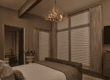 A bedroom with a bed, textured bedding, an antler-style chandelier, large windows with drapes, and a doorway leading to a bathroom with a tub and a vase of flowers on a small table.