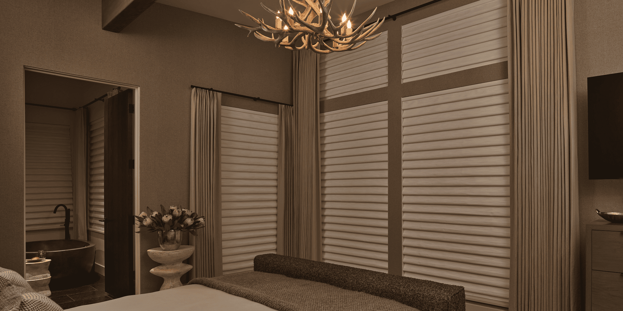 2023 AURA VIG PV Belfast Linen Bed Down Night (1) A bedroom with a bed, textured bedding, an antler-style chandelier, large windows with drapes, and a doorway leading to a bathroom with a tub and a vase of flowers on a small table.