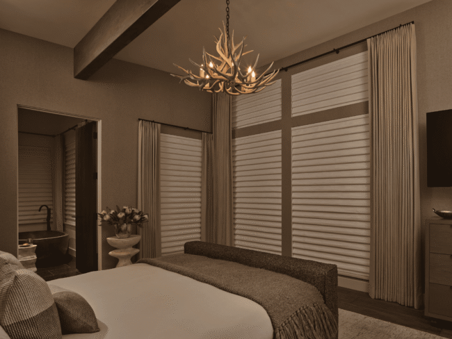 A bedroom with a bed, textured bedding, an antler-style chandelier, large windows with drapes, and a doorway leading to a bathroom with a tub and a vase of flowers on a small table.