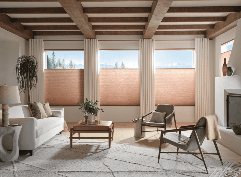 Modern living room with a white sofa, two chairs, a coffee table, large windows with beige shades, wooden beams on the ceiling, a fireplace, and neutral decor elements.