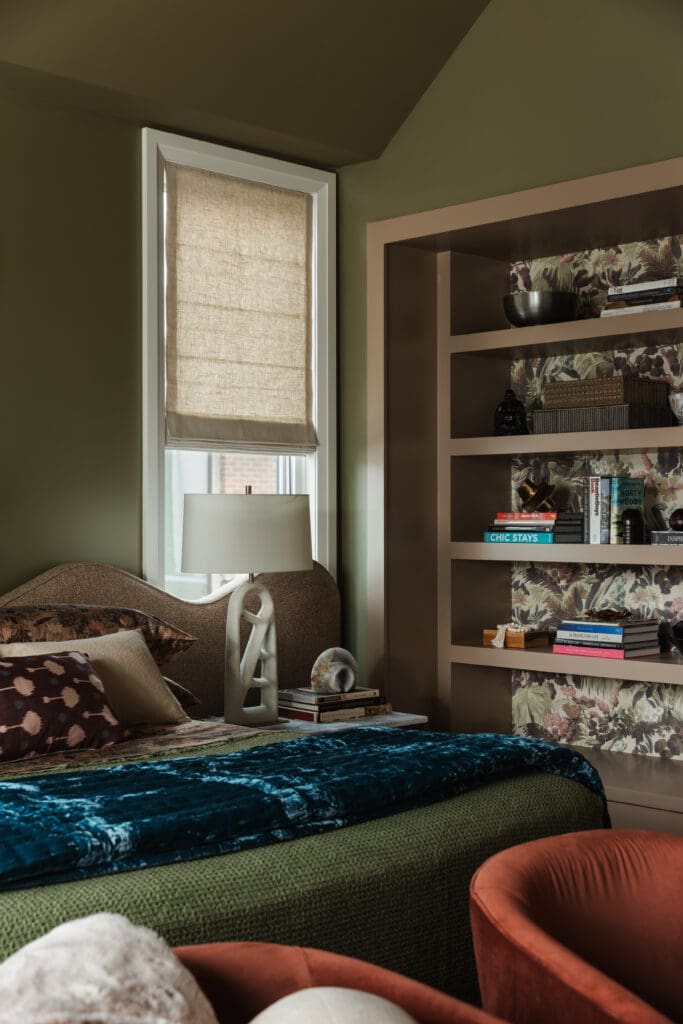 A cozy bedroom with a made bed, abstract pillows, lamp, books on a side table, a window with a shade, and a built-in bookshelf filled with books and decorative items.
