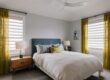 A modern bedroom with a blue upholstered bed, white bedding, two wooden nightstands with lamps, yellow curtains, and two large windows.